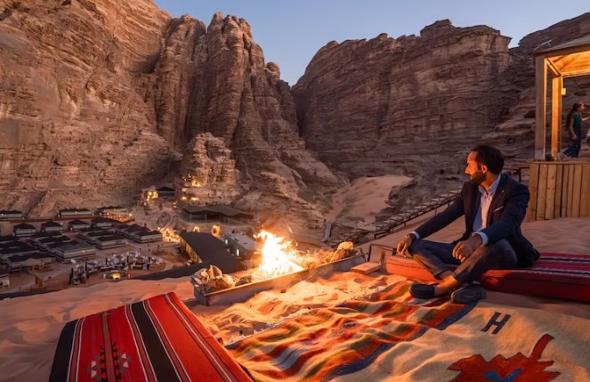 Man sitting above a Bedouin desert camp in Wadi Rum Jordan at sunset with sandstone cliffs firelight and dramatic mountain scenery