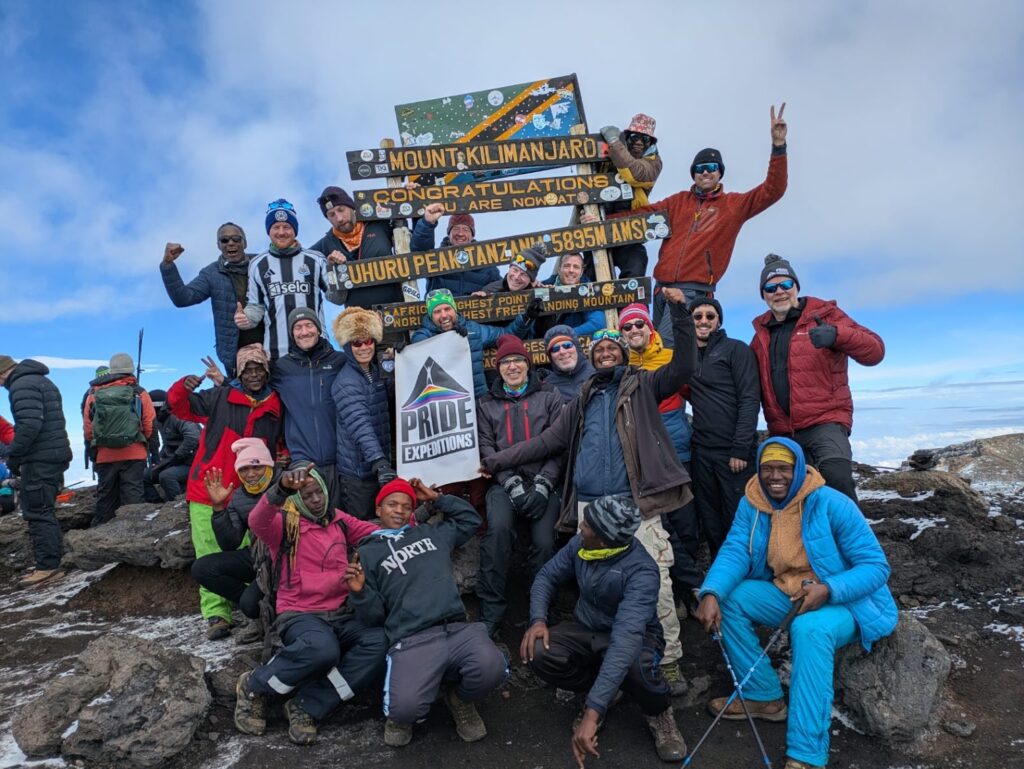 Pride Adventures and Pride Expeditions climbers with mountain guides at Uhuru Peak summit sign on Mount Kilimanjaro, Tanzania.