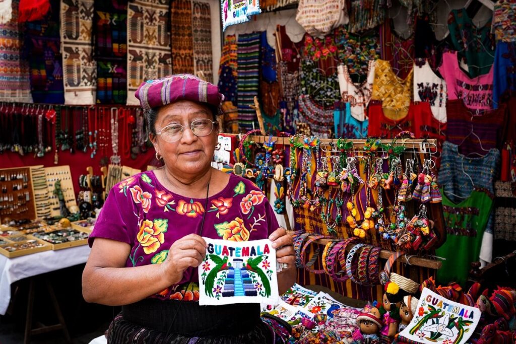 Maya woman in traditional purple huipil displaying colorful Guatemala souvenir textiles at Chichicastenango market