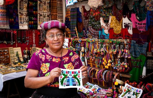 Maya woman in traditional purple huipil displaying colorful Guatemala souvenir textiles at Chichicastenango market