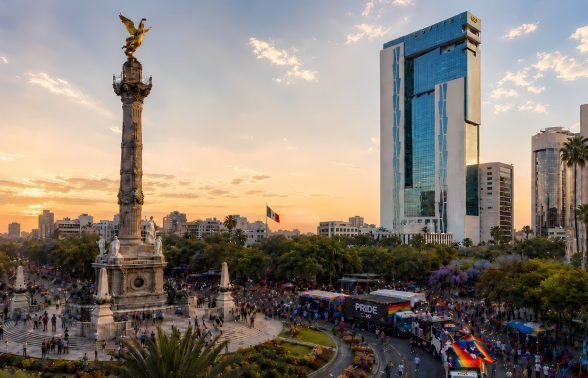 Golden hour aerial view of Mexico City with the Angel of Independence monument, Sofitel hotel, and Pride festival activities below at sunset