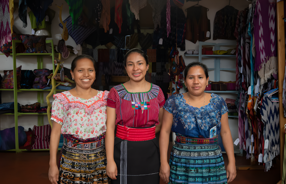 Three Tz'utujil Maya women in vibrant traditional woven dress smiling inside a textile shop in Guatemala